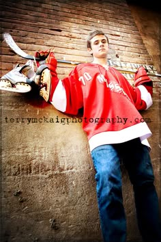 a young man leaning against a brick wall wearing a red hockey jersey and holding a pair of skis