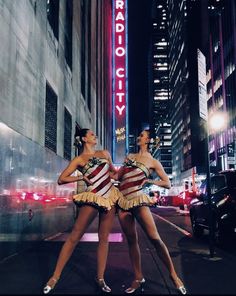 two women dressed in costumes standing on the side of a city street at night with neon signs behind them