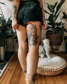 a woman with tattoos on her legs standing in front of a cat and potted plant