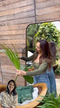 two women standing in front of a wooden wall with plants on it and one woman holding a palm leaf