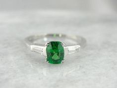 The color combination of this one of a kind engagement ring is eye catching! We've set a perfect, leaf green Tsavorite Garnet in the center of this vintage mounting, creating a piece that is unique and one of a kind. Accented with tapering, baguette diamonds, this is a great piece for every day or special occasions. We've set this ring with a new, bright green Tsavorite Garnet. Mined in Africa, Tsavorite was named for Tsavo National Park, in Kenya. The green shade is slightly brighter, a little Art Deco Green Ring, Gia Certified, Art Deco Green Ring Gia Certified, Vintage Green Emerald Ring Gia Certified, Gia Certified Green Diamond Ring In Vintage Style, Vintage Green Emerald Ring With Center Stone, Heirloom Style Green Emerald Ring With Center Stone, Heirloom Green Emerald Ring With Center Stone, Vintage Green Gia Certified Rings, Green Diamond Heirloom Ring With Center Stone