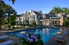 an outdoor swimming pool surrounded by lawn furniture and flowers in front of a large house