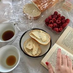 a person reading a book with bread, raspberries and coffee on the table