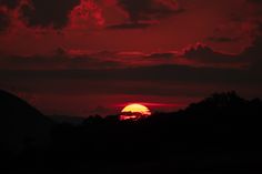 the sun is setting over some hills with trees in front of it and red clouds