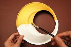 two hands painting a yellow and white circle with paintbrushes on a brown surface