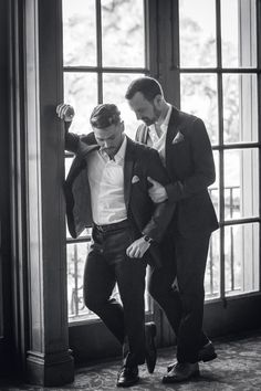 two men standing next to each other in front of a window wearing suits and ties