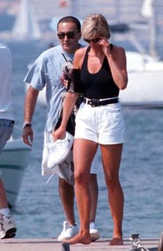 a man and woman walking on a dock next to the water with boats in the background