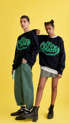 two people standing next to each other in front of a yellow background wearing black and green sweatshirts