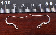 two silver hooks are sitting next to a measuring ruler on a wooden surface, with one hook in the middle