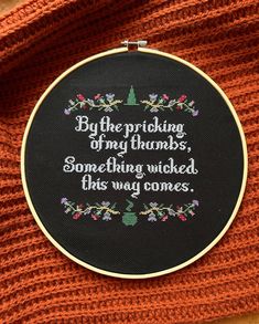 a cross stitched quote on a black hoop hanging from an orange sweater with the words, by the pricking of my thumb, something, something i'd