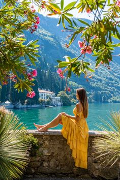 a woman in yellow dress sitting on stone wall next to water with mountains and flowers