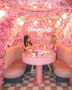 two women sitting at a pink table with flowers on the wall