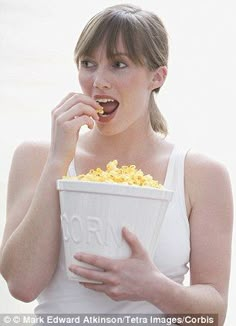 a woman is eating popcorn out of a white bucket while standing on the beach with her hands in her mouth