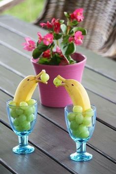 two glasses with grapes and bananas in them on a wooden table next to a potted plant