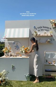 a woman standing in front of a white building with flowers on the wall and shelves