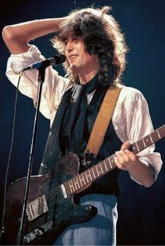 a man with long hair holding a guitar in front of a microphone and wearing a white shirt