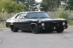 an old black muscle car parked in a parking lot next to some trees and bushes