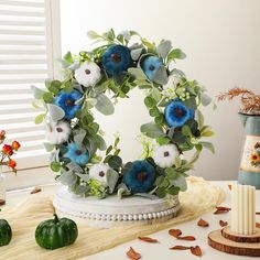 a wreath with blue flowers and greenery on a table next to candles, pumpkins and other decorations