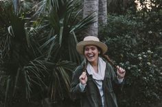 a woman in a hat is smiling and holding her hands up while walking through the woods