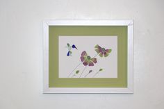 a white frame with some purple flowers and green leaves on it, hanging on the wall