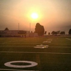 the sun is setting over a football field with white numbers painted on it and trees in the background