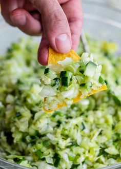 a hand holding a tortilla chip over a bowl of shredded cabbage and cucumbers