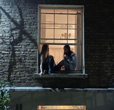 two people sitting on the ledge of a building at night looking out an open window