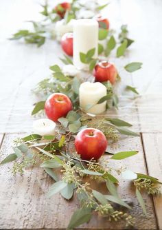 an arrangement of apples and greenery on a wooden table with candles in the background