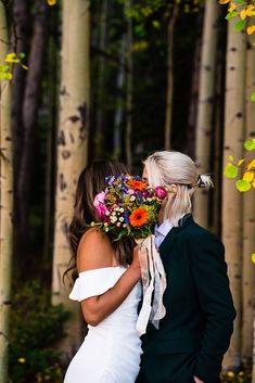 a bride and groom standing in front of trees with flowers on their wedding bouquets