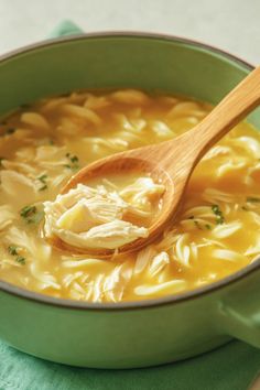 a wooden spoon in a green pot filled with chicken noodle soup on a table