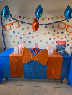 an orange and blue birthday party table with balloons, streamers and bags on it