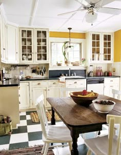 a kitchen filled with lots of white furniture and yellow wall paint on the walls, along with a wooden table topped with bowls of fruit