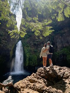 two people standing on top of a rock next to a waterfall and looking at the water