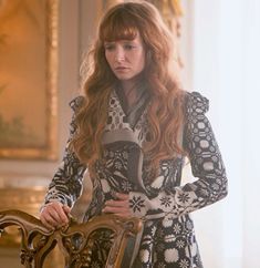 a woman with long red hair standing next to a chair and looking at the camera