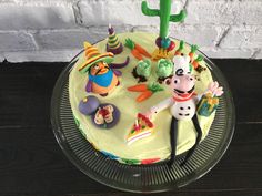 a birthday cake decorated with toy figures and decorations on a glass platter next to a brick wall