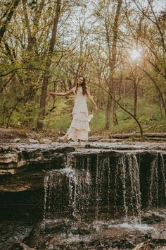 a woman in white dress standing on top of a waterfall with her arms spread out