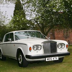 an old white car parked on the grass in front of a brick building and trees