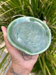 a hand holding a green bowl with designs on the outside and inside, in front of some plants