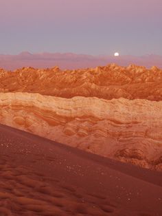 the moon shines brightly in the distance over desert terrain, with mountains and sand dunes