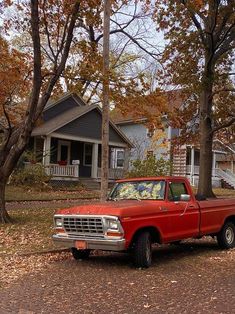 an old red pickup truck parked in front of a house surrounded by trees and leaves