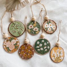 six hand painted wooden ornaments hanging from strings on a white cloth covered table with flowers and leaves