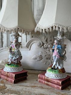 two figurines sitting on top of books in front of a lampshade