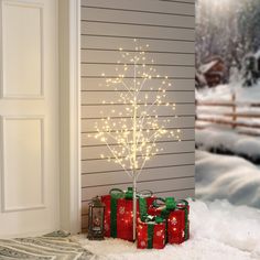 a small christmas tree with lights in front of a house and presents on the snow