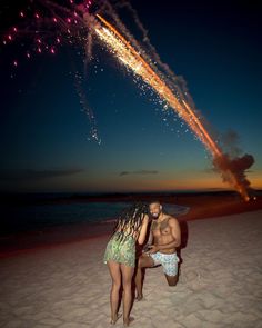 two people on the beach with fireworks in the sky above them and one person kneeling down