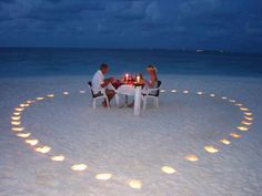 two people sitting at a table on the beach with candles in the shape of a heart