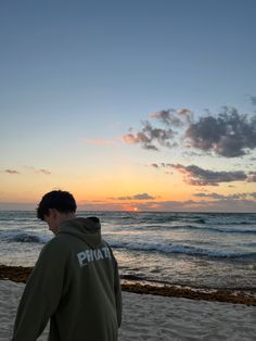 a young man standing on top of a sandy beach next to the ocean at sunset