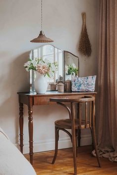 a desk with a mirror, vase and flowers on it in front of a window