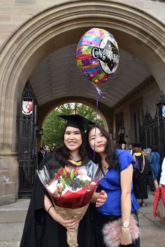 two women standing next to each other in graduation gowns holding flowers and balloons with confetti on them