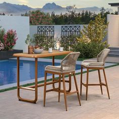 two wicker bar stools sitting next to a table near a swimming pool with mountains in the background