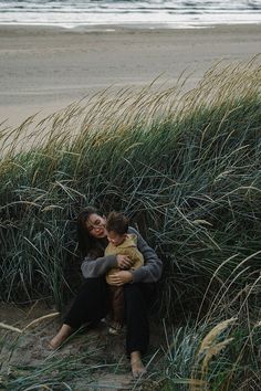 a woman holding a child sitting on top of a grass covered field next to the ocean